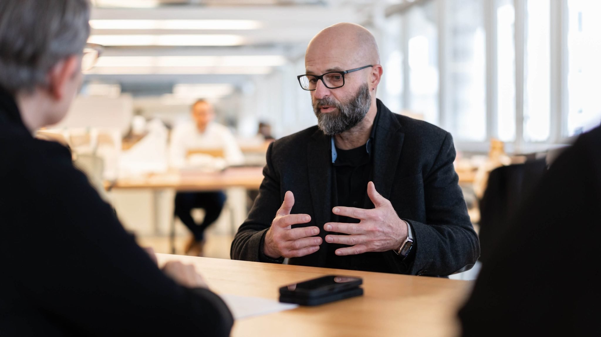 A horizontal, close-up shot of a man with a beard and a shaved head, wearing glasses. He is sitting at a wooden table and leaning forward, gesturing with both hands as he speaks. The person he is speaking to is visible on the far left of the frame, facing away from the camera. The background is a large, well-lit, open-plan office space with tables and chairs.
