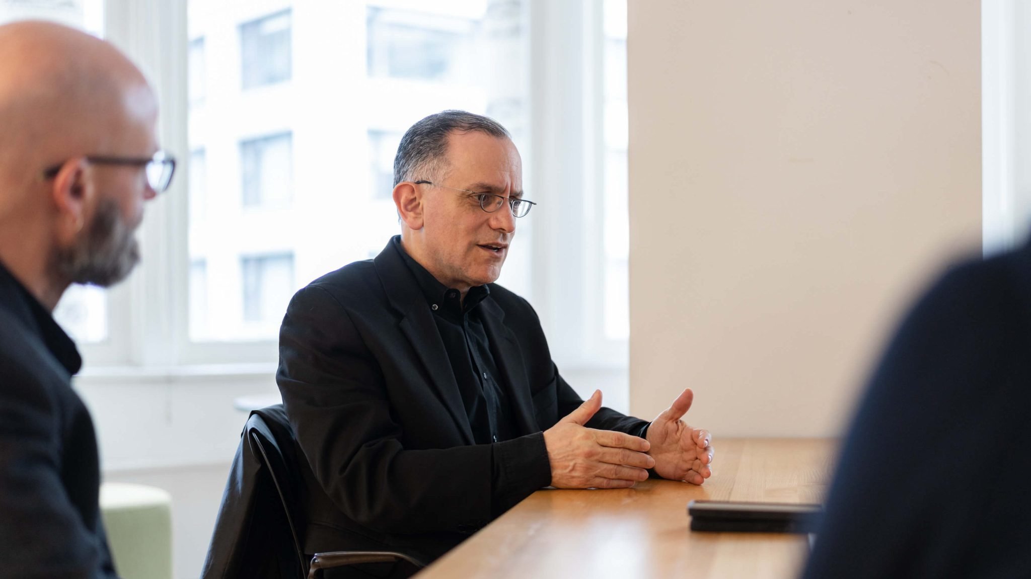 A horizontal, close-up shot of a man with short hair and glasses. He is sitting at a light wooden table and is wearing a black shirt and a black jacket. He is looking to his left and speaking, with both hands on the table gesturing. The person he is speaking to is visible on the far left of the frame, facing away from the camera. The background is a white wall with a window.