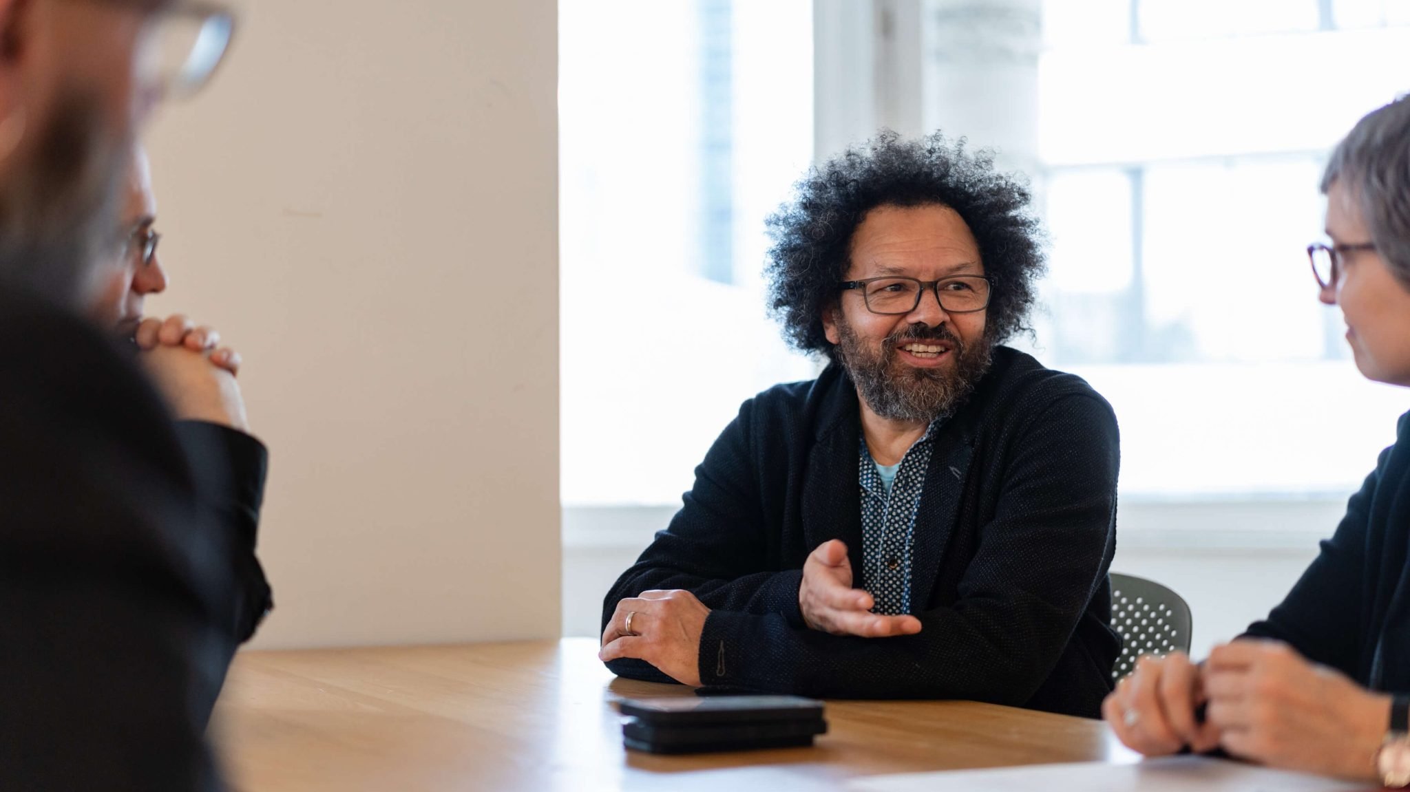 A horizontal, close-up shot of three people sitting at a wooden table in a well-lit room. The central figure is a Black man with a beard, curly hair, and glasses, wearing a dark blue shirt and a dark cardigan. He is smiling and gesturing with his right hand as he talks to the person on his right, whose back is to the camera. The person on the left is only partially visible and is also looking toward the central figure.